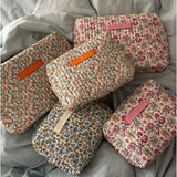 Fem blomstrede toilettasker med mærket "LA PETITE CACHOTIERE" ligger på et lyseblåt dynebetræk. Taskerne har forskellige farver blomster, og mærkerne har orange, lyserød eller beige baggrund.