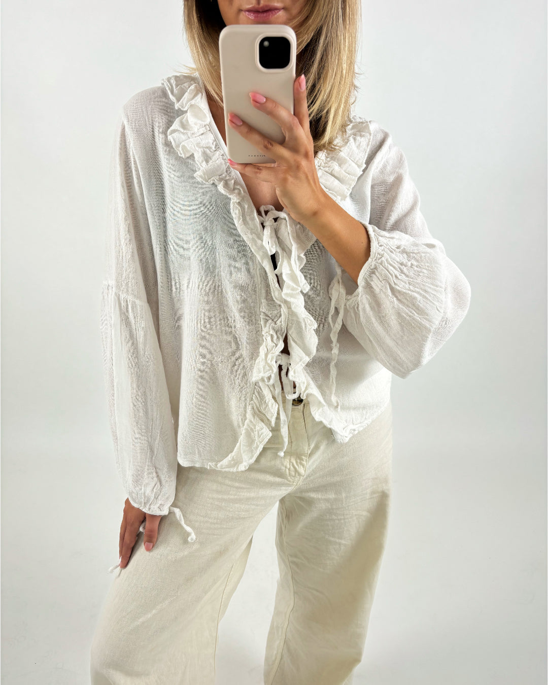 En kvinde tager et selfie med en beige telefon. Hun er iført en hvid, løstsiddende bluse med flæser og beige bukser i en hvid baggrund.