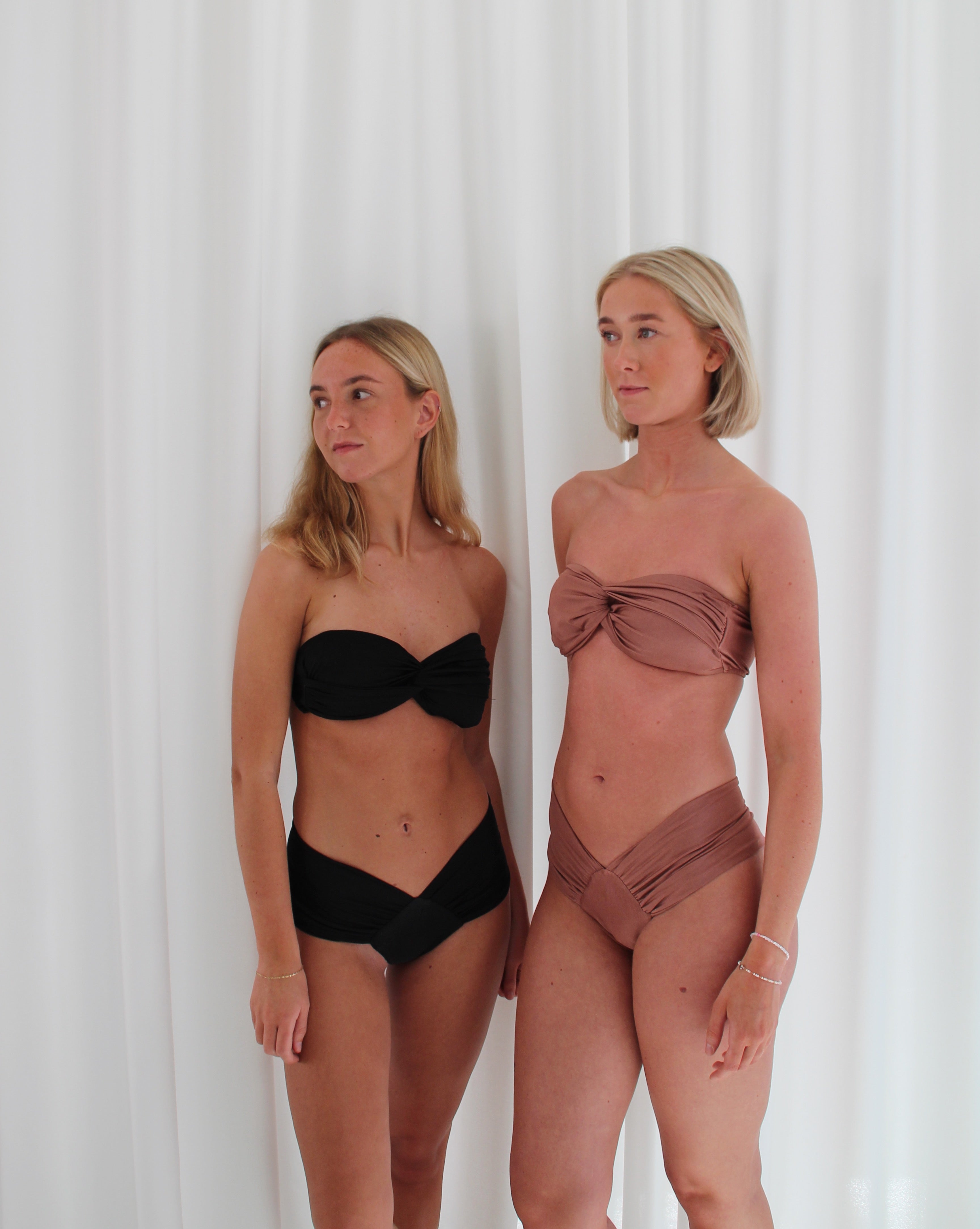 To blonde kvinder poserer i matchende bikinis foran et hvidt lagen. Den ene bærer en sort bikini, mens den anden bærer en mokkafarvet.
