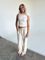 En kvinde står og smiler, iført en hvid top og løse, beige bukser med snøre i taljen, i et lyst studiemiljø.