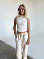 En person står med hænderne i lommerne, iført en hvid top og løse lyse bukser, i et minimalistisk rum med grå gulv og hvide vægge.