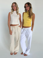 To kvinder står side om side iført lette, ærmeløse toppe og løse bukser. Den ene top er hvid, den anden gul. De befinder sig i et minimalistisk, lyst rum.