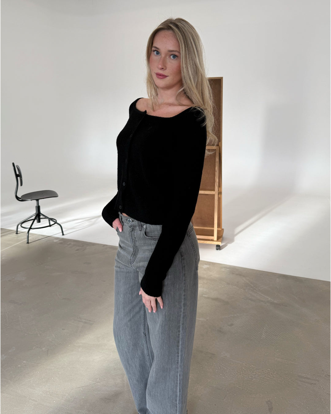 En person står afslappet iført en sort trøje og grå jeans. De befinder sig i et minimalistisk rum med en stol og en trækasse i baggrunden.