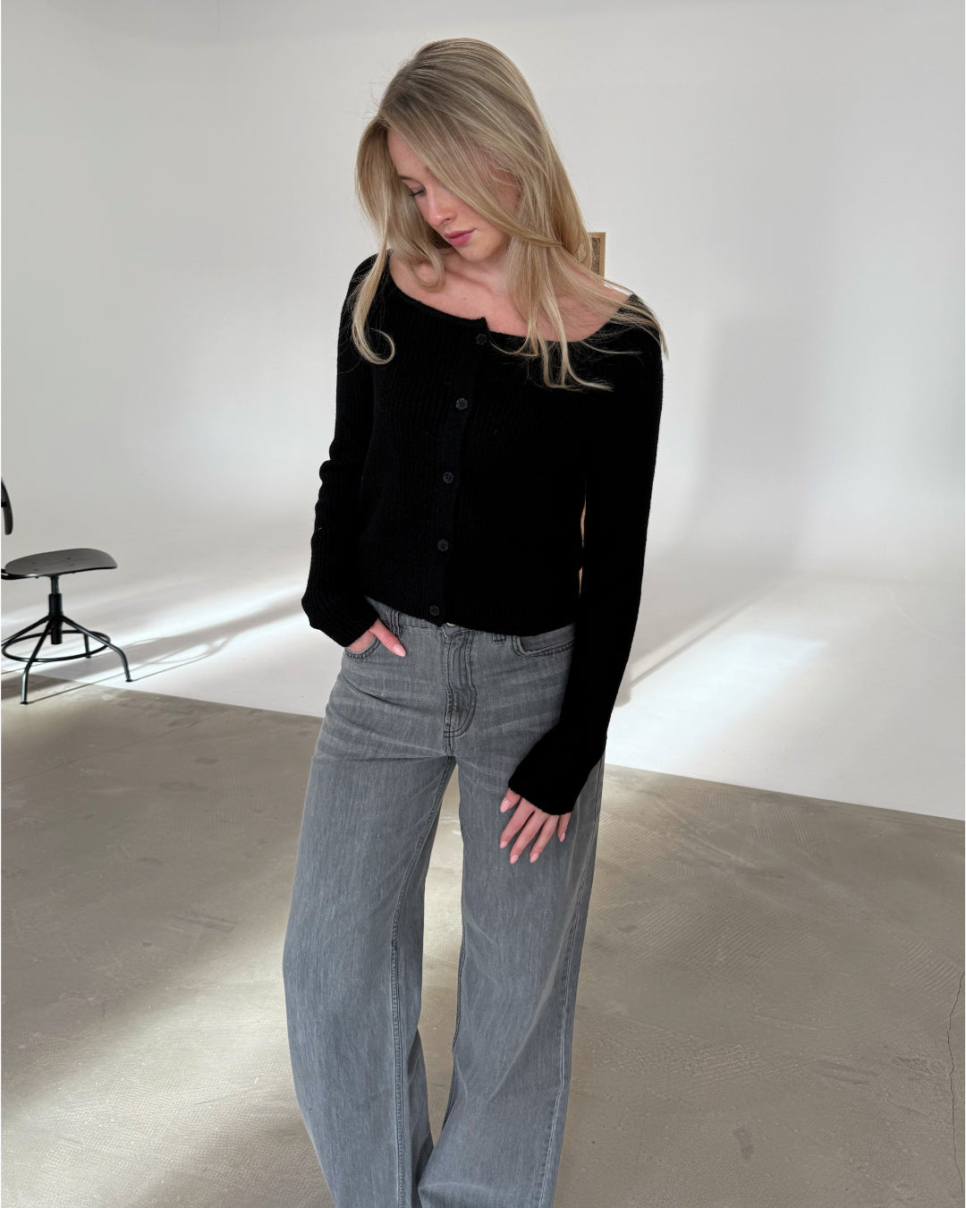 Kvinde ser ned, iført sort trøje og grå jeans, i et minimalistisk rum med lys væg og en sort stol.