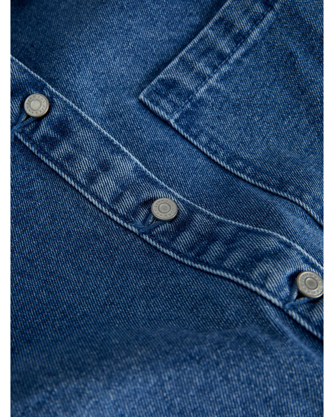 Denimstof med tre sølvknapper i en diagonal linje, del af en skjorte eller jakke, der ligger fladt oplyst i et neutralt lys.