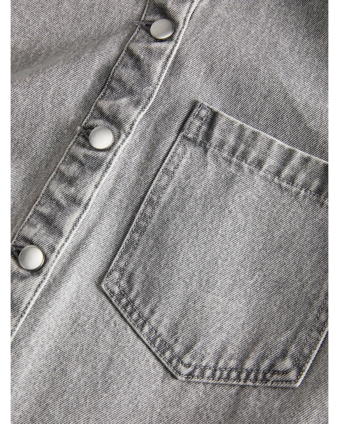 Brystlomme placeret på en grå denimskjorte med sølvfarvede trykknapper. Stoffets tekstur er tydelig i nærbilledet.