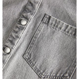 Brystlomme placeret på en grå denimskjorte med sølvfarvede trykknapper. Stoffets tekstur er tydelig i nærbilledet.