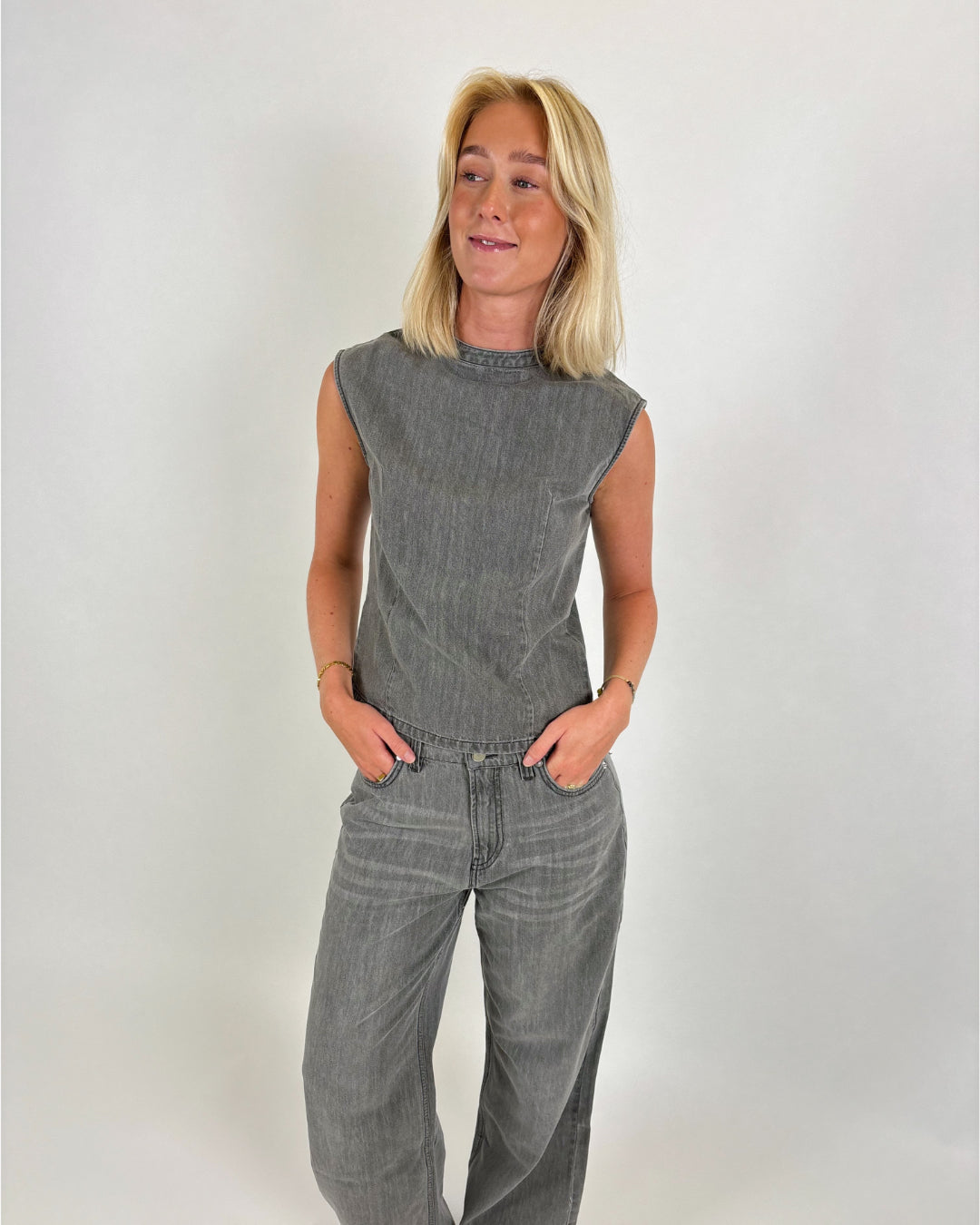 En person i gråt denim tøj står med hænderne i lommerne, ser til siden og smiler let i et neutralt studieomgivelser.