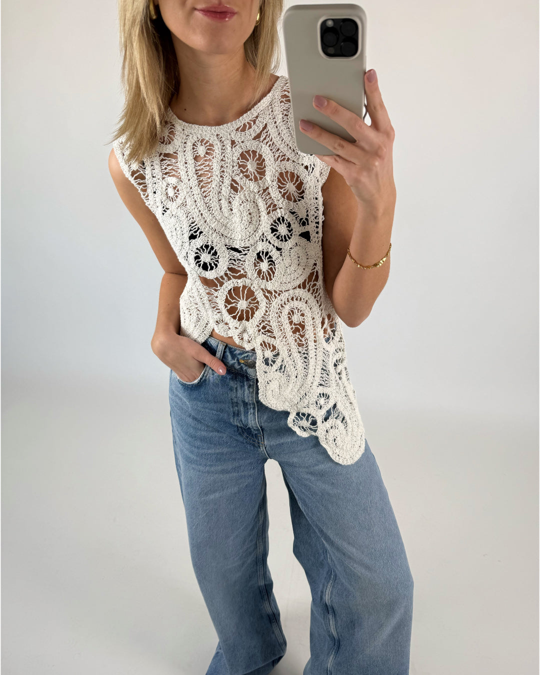 En person iført en hvid, detaljeret hæklet top tager en selfie med en smartphone. Personen har også lyseblå jeans på og står i et minimalistisk rum med lyse vægge.