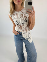 En person iført en hvid, detaljeret hæklet top tager en selfie med en smartphone. Personen har også lyseblå jeans på og står i et minimalistisk rum med lyse vægge.