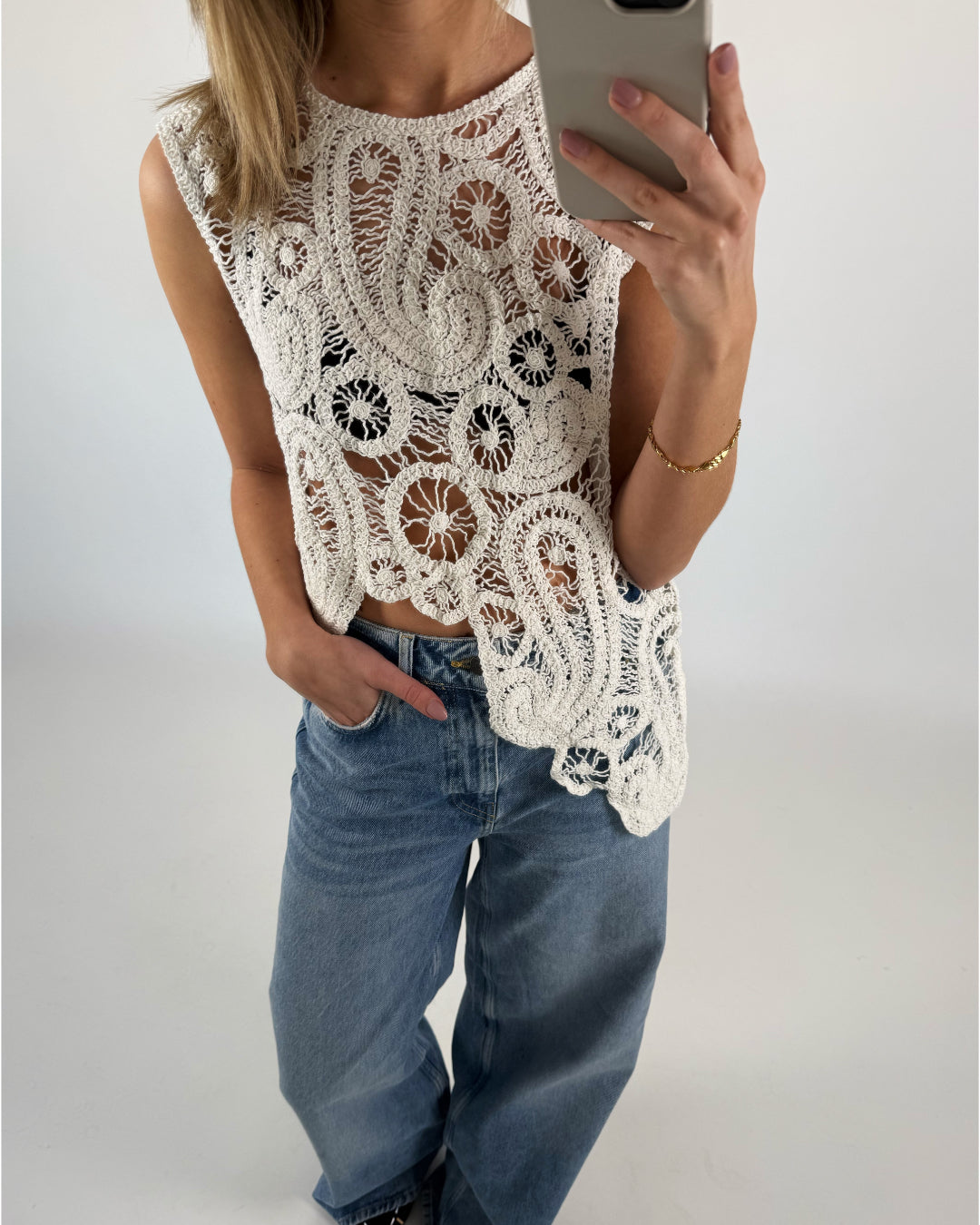 En person iført en hvid, mønstret, hæklet top tager et spejl-selfie og har en hånd i lommen på et par blå jeans i et neutralt indendørsmiljø.