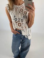 En person iført en hvid, mønstret, hæklet top tager et spejl-selfie og har en hånd i lommen på et par blå jeans i et neutralt indendørsmiljø.