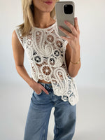 En kvinde iført en hvid, mønstret, ærmeløs top tager et selfie med en smartphone. Hun står i lyseblå jeans i et minimalistisk rum.