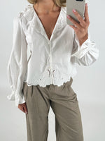 En person iført en hvid bluse med broderede detaljer tager en selfie med en smartphone i et neutralt indendørs miljø. Personen har også brune bukser på.