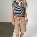 En person står iført en sort- og hvidstribet trøje og beige shorts. De står på et gulv med to forskellige farver, brun og hvid, i en enkel, lys tekstur.