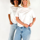 To personer i hvide T-shirts og jeans står smilende i et studie med hvid baggrund. Den ene person har krøllet hår, den anden har skulderlangt blond hår.