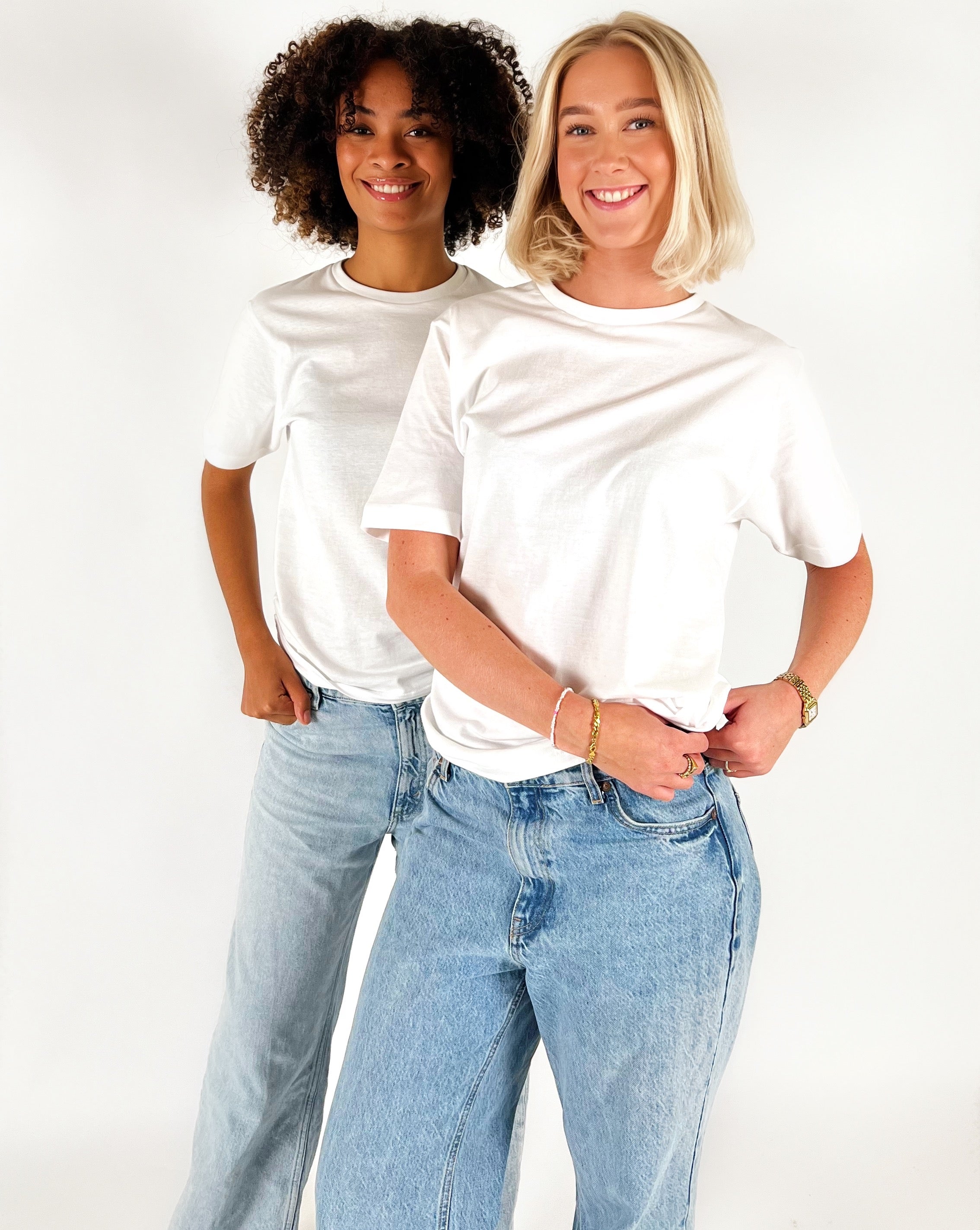 To personer i hvide T-shirts og jeans står smilende i et studie med hvid baggrund. Den ene person har krøllet hår, den anden har skulderlangt blond hår.