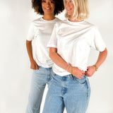 To kvinder iført hvide T-shirts og blå jeans står ved siden af hinanden og smiler i et lyst studiemiljø.