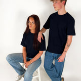 En person sidder på en hvid stol, iført en sort t-shirt og jeans, mens en anden person står ved siden af og hviler en arm på stolens ryglæn. Begge er i et lyst rum.