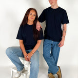 To personer i sorte t-shirts og blå jeans; den ene sidder på en hvid skammel, mens den anden står ved siden af i et lyst rum.