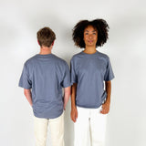 To personer står ved siden af hinanden; en vender ryggen til, mens den anden ser fremad. De bærer begge grå t-shirts og hvide bukser, i et neutralt rum.