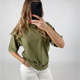 Kvinde tager en selfie med en sort telefon. Hun har en grøn t-shirt og hvide bukser på, i et neutralt oplyst rum. Hendes negle er rødlakkerede.