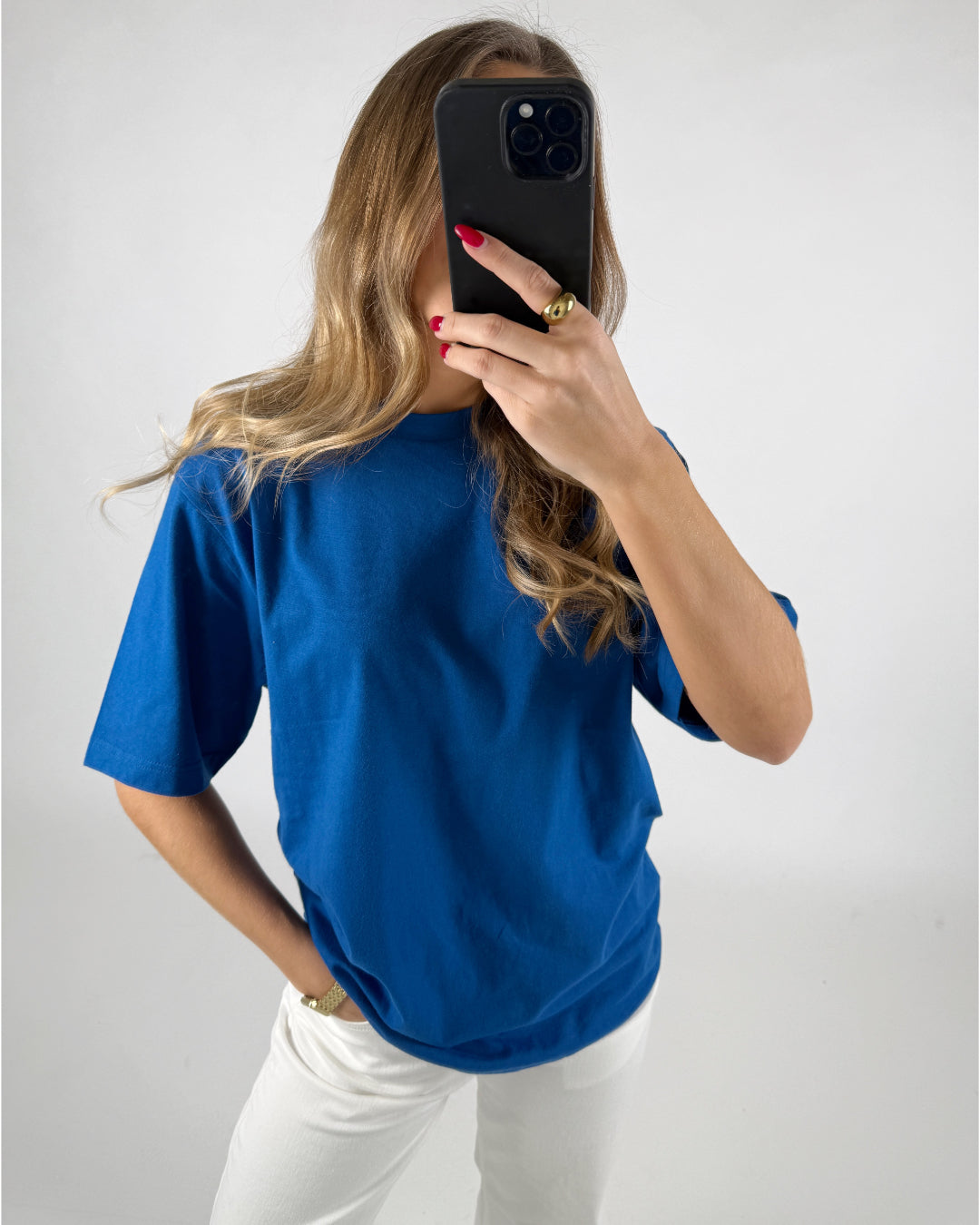 En person i en blå t-shirt holder en smartphone op foran ansigtet i en selfie-position. Personen står i et lyst rum med hvid baggrund.