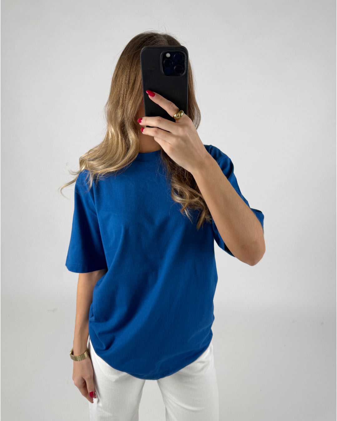 En person med langt hår står i en blå t-shirt og hvide bukser og holder en sort telefon op foran ansigtet i et lysomt rum.