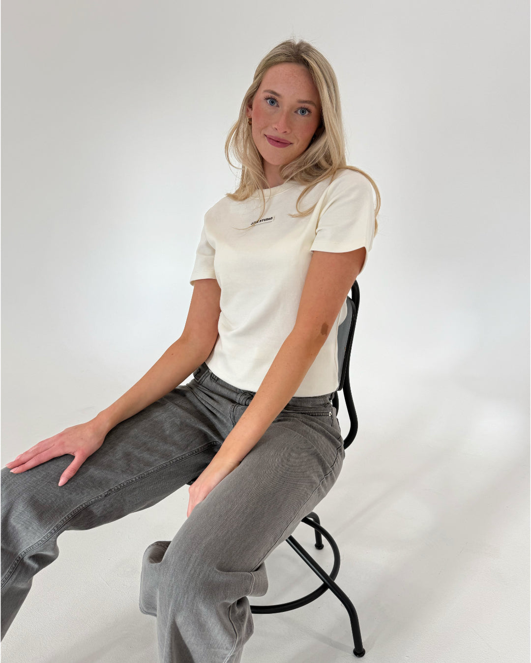 En kvinde sidder afslappet på en stol, iklædt en hvid T-shirt og grå bukser, i et lyst rum uden synlig indretning.