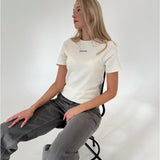 En person sidder på en stol iført en hvid t-shirt med teksten "ASOS DESIGN" og grå jeans. Baggrunden er en simpel, lys væg.