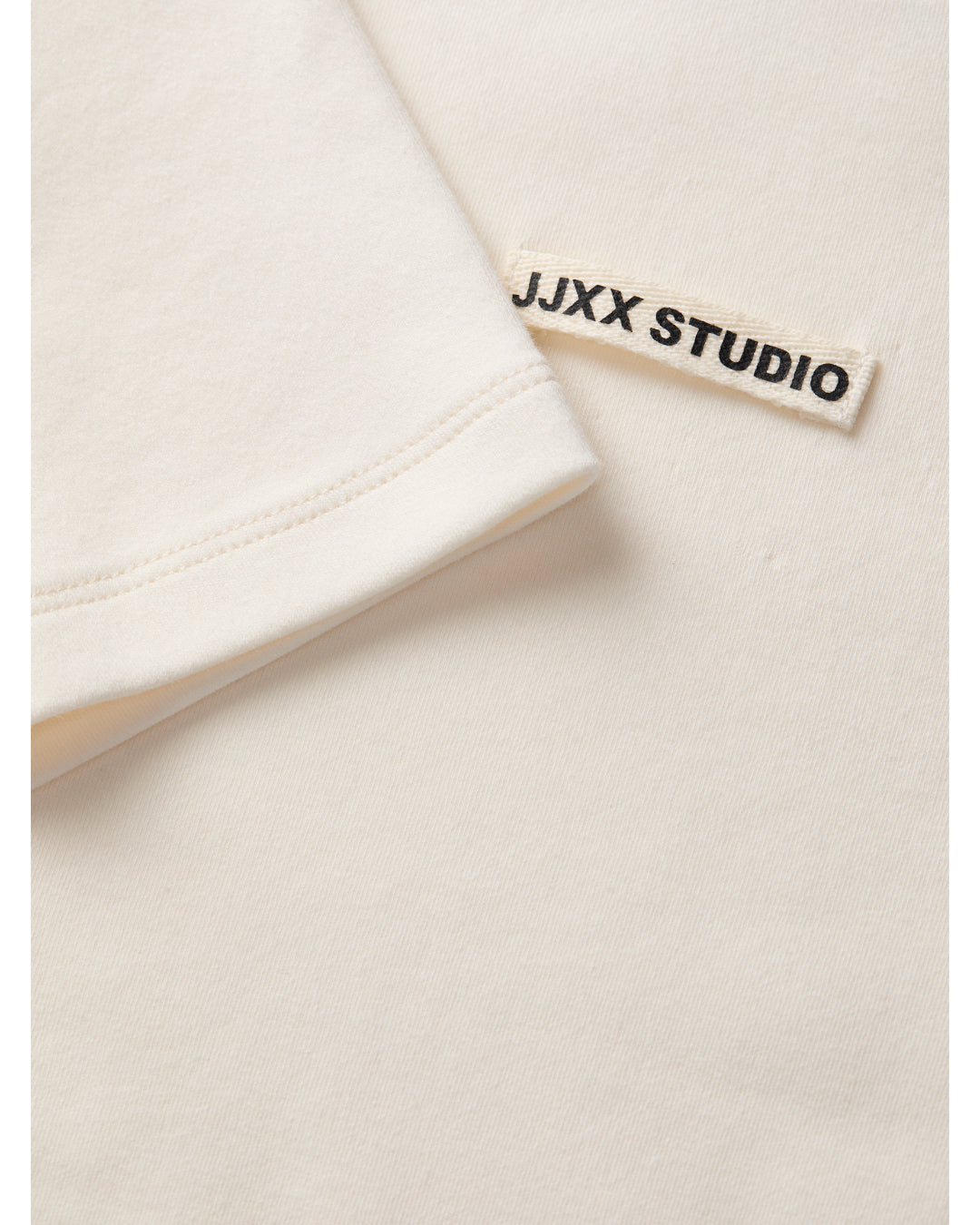 En hvid T-shirt har en påsyet etikette med teksten "JJXX STUDIO" i sorte bogstaver, set i en lys, ensartet baggrund.