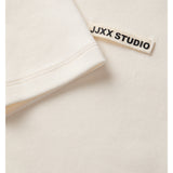 En hvid T-shirt har en påsyet etikette med teksten "JJXX STUDIO" i sorte bogstaver, set i en lys, ensartet baggrund.