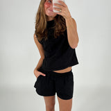 En kvinde iført sort top og shorts tager en selfie med en smartphone i et hvidt studie. Hun smiler og har hånden i lommen.