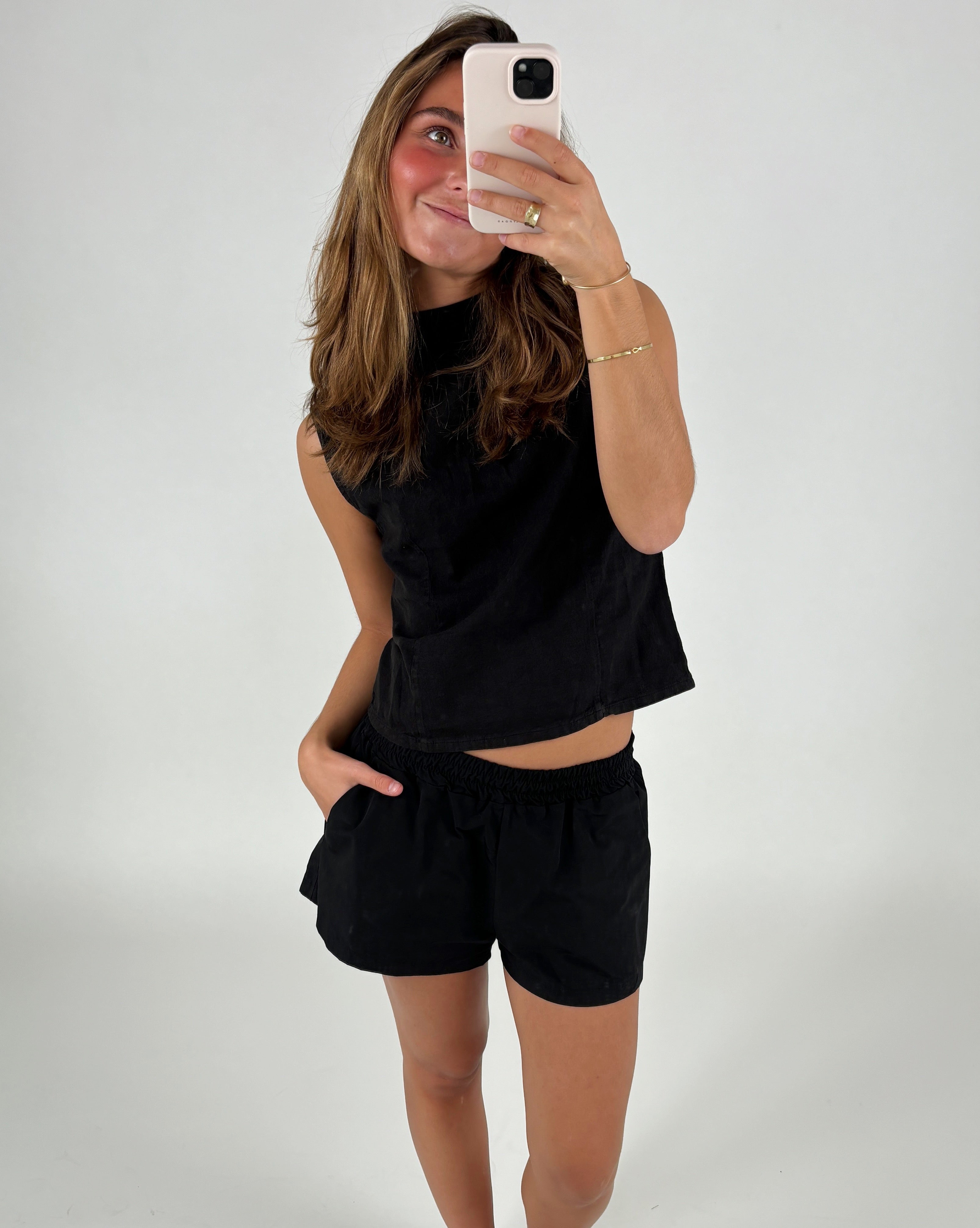 En kvinde iført sort top og shorts tager en selfie med en smartphone i et hvidt studie. Hun smiler og har hånden i lommen.