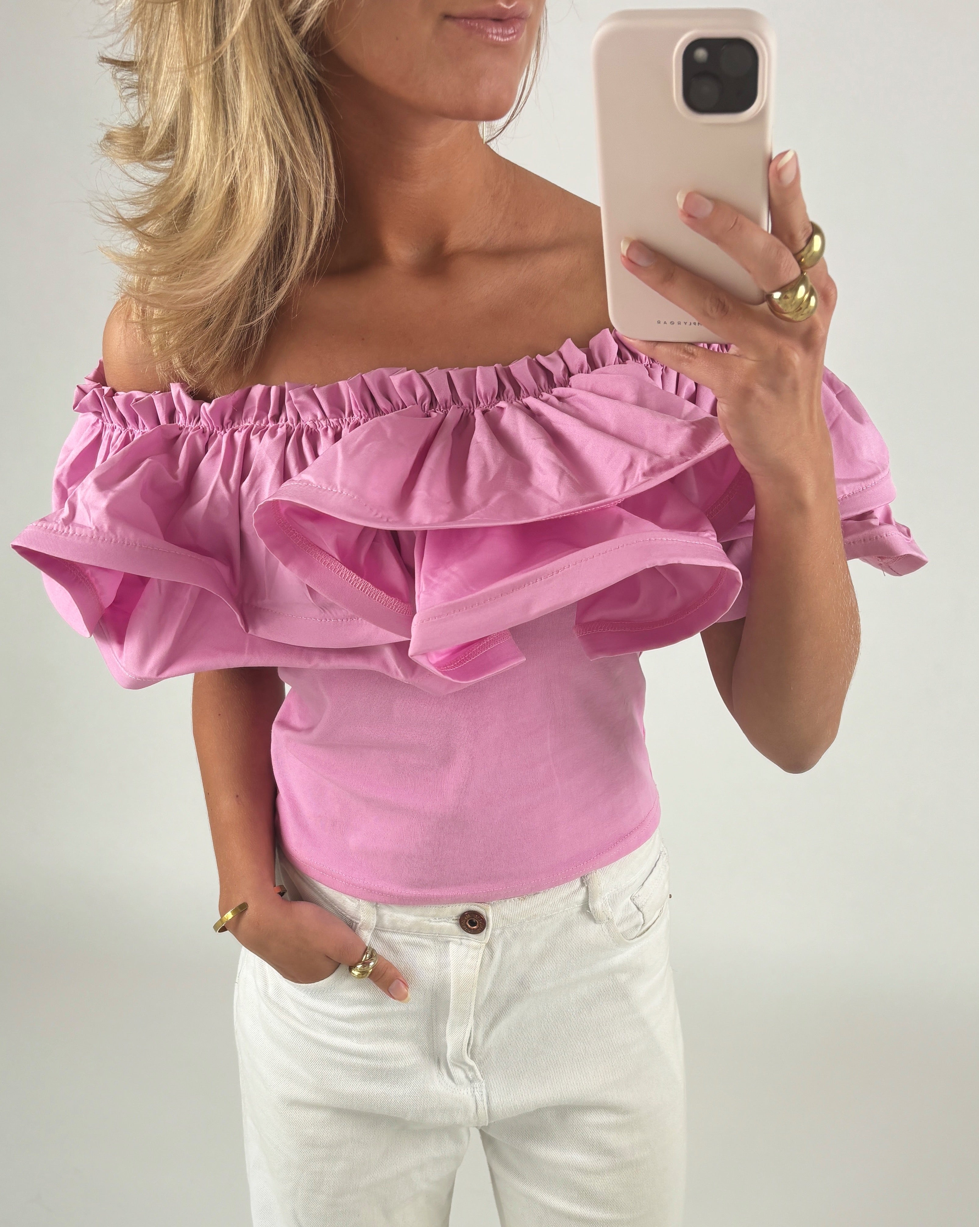En person iført en pink, off-shoulder top og hvide bukser holder en smartphone og tager et billede i et lyst rum. Personen har blondt hår og bærer guldsmykker.