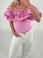 En kvinde iført en lyserød, skulderfri top med flæser står med hånden i lommen på hvide bukser. Baggrunden er enkel og ensfarvet.