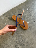 En hånd holder en brun sandal med blå stropper, mens den ligger på en grå betonoverflade ved siden af en hvid væg.