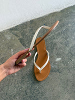 En hånd holder en brun og hvid flip-flop-sandal. Baggrunden er et råt, gråt betonunderlag ved en hvid væg.