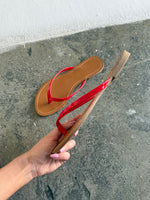 En flip-flop med røde remme holdes i en hånd mod en ru, grå cementbaggrund nær en hvid væg. En anden lignende sandal ligger på gulvet.