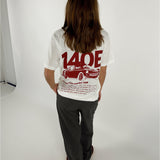 En person står med ryggen til iført en hvid T-shirt med rød grafik af en bil og tekst "140E Racing Club member 140E ... 1987 ... Car racing ...". De er i en lys indendørs omgivelser.