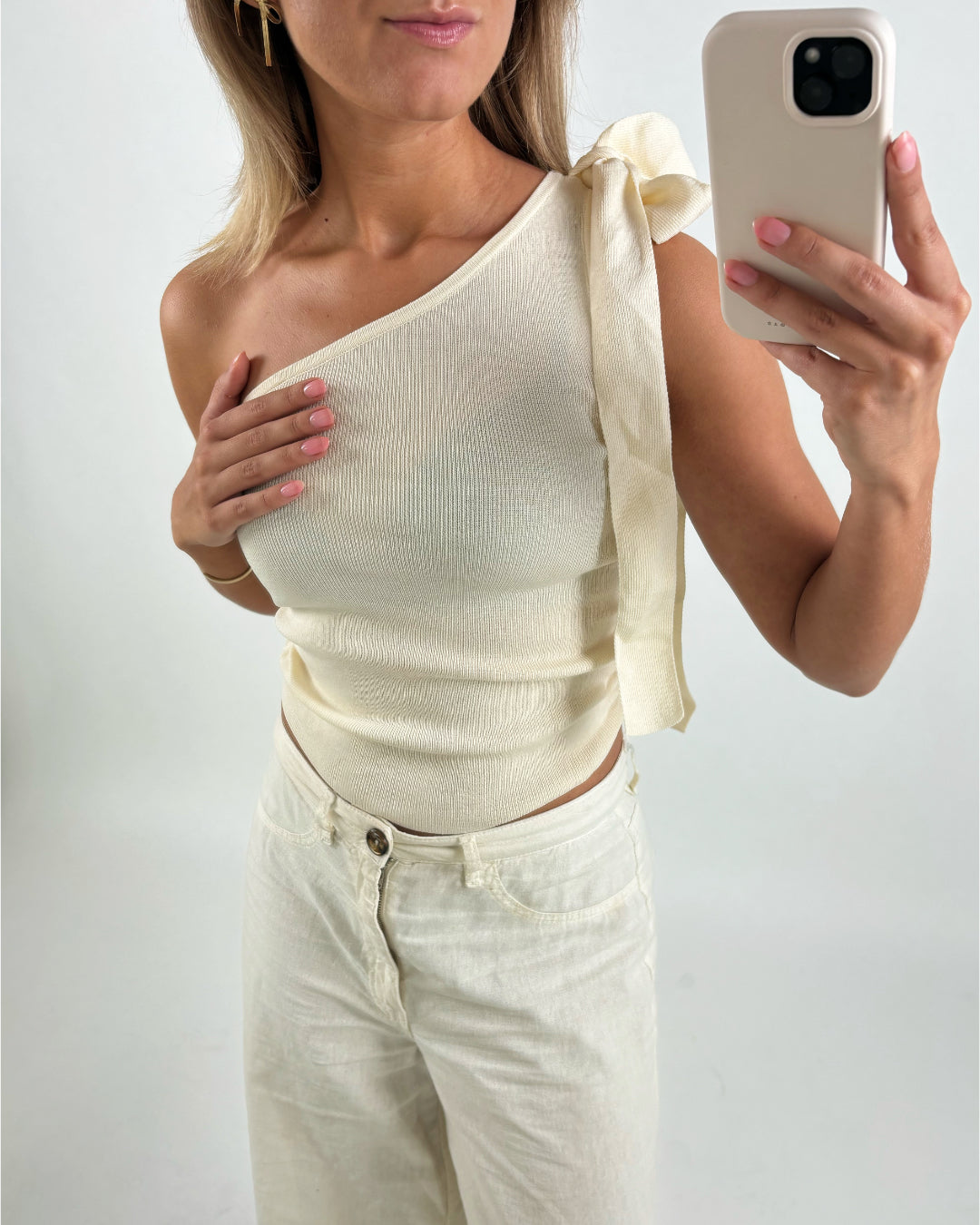 En person iført et off-shoulder, cremefarvet top med en sløjfe holder en mobiltelefon foran et hvidt baggrund. Personen har cremefarvede bukser på og kort lyst hår.