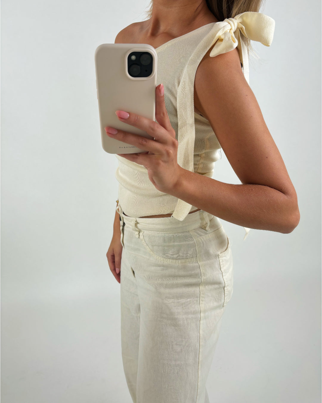 En person iført en cremefarvet top med skulderbånd holder en mobiltelefon op foran et hvidt baggrundsmiljø. Personen er også klædt i matchende bukser.