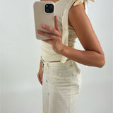 En person iført en cremefarvet top med skulderbånd holder en mobiltelefon op foran et hvidt baggrundsmiljø. Personen er også klædt i matchende bukser.