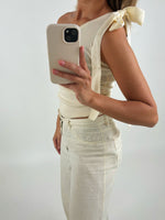 En person iført en cremefarvet top med skulderbånd holder en mobiltelefon op foran et hvidt baggrundsmiljø. Personen er også klædt i matchende bukser.