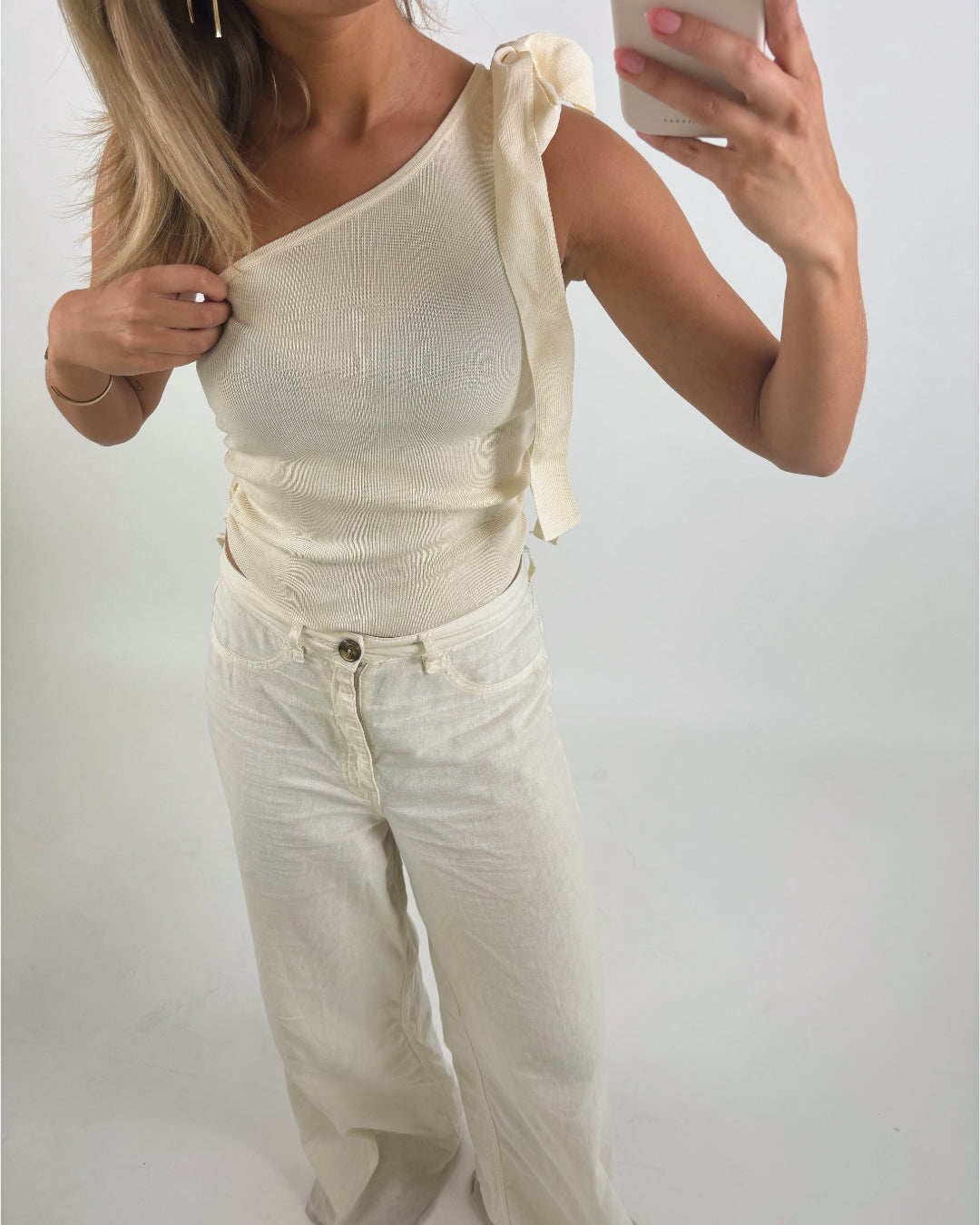 Kvinde i beige top med ene skulder op i knude, bærer cremefarvede bukser, tager selfie foran en neutral baggrund. Hun holder en mobiltelefon i venstre hånd.