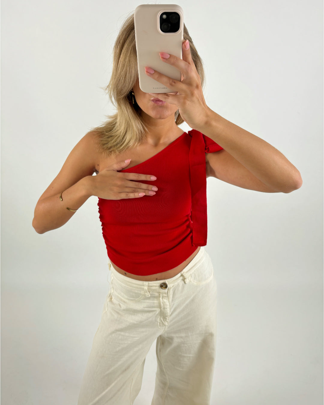 Person tager et spejlbillede med en smartphone, iført en rød top og hvide bukser, i et rum med lys baggrund. Hånden dækker delvist brystet.