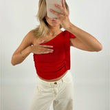 Person tager et spejlbillede med en smartphone, iført en rød top og hvide bukser, i et rum med lys baggrund. Hånden dækker delvist brystet.