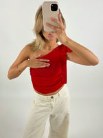 Person tager et spejlbillede med en smartphone, iført en rød top og hvide bukser, i et rum med lys baggrund. Hånden dækker delvist brystet.