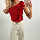 Kvinde i rød, ærmeløs top tager en selfie med telefon i højre hånd, iført hvide bukser, mod en neutral baggrund. Hun retter på sin top med venstre hånd.