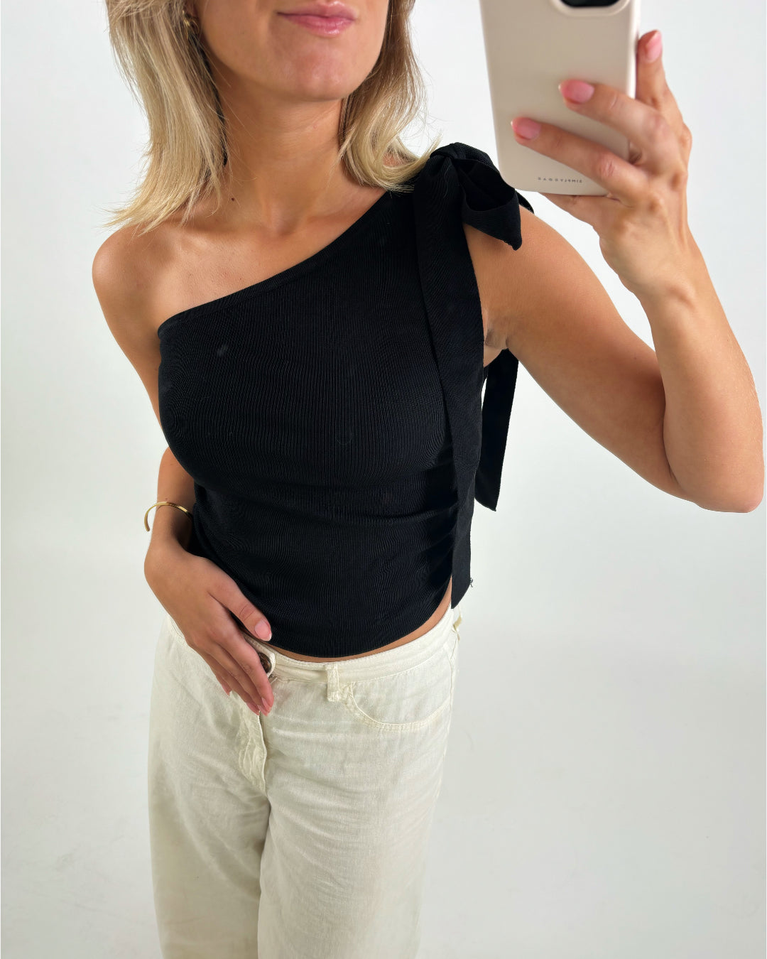 Person tager selfie iført sort, skulderfri top og lyse bukser, med mobilen i hånden. De står i et lyst rum, og telefonens mærke "CASEYNI" er synligt.